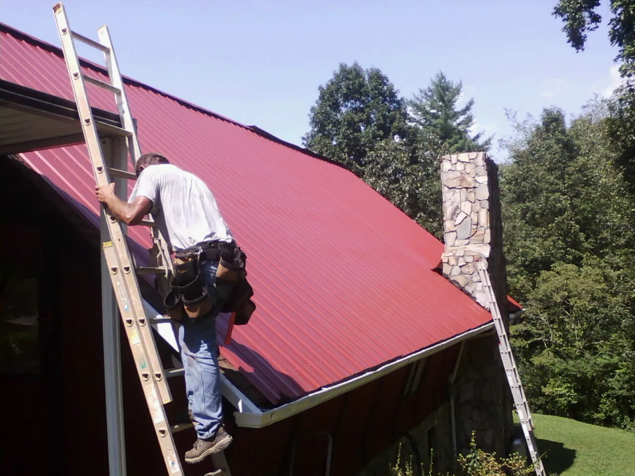 Roofer on a ladder beside a red metal roof and stone chimney — Soffit & Fascia Repair by Roofing Brothers in Willamina